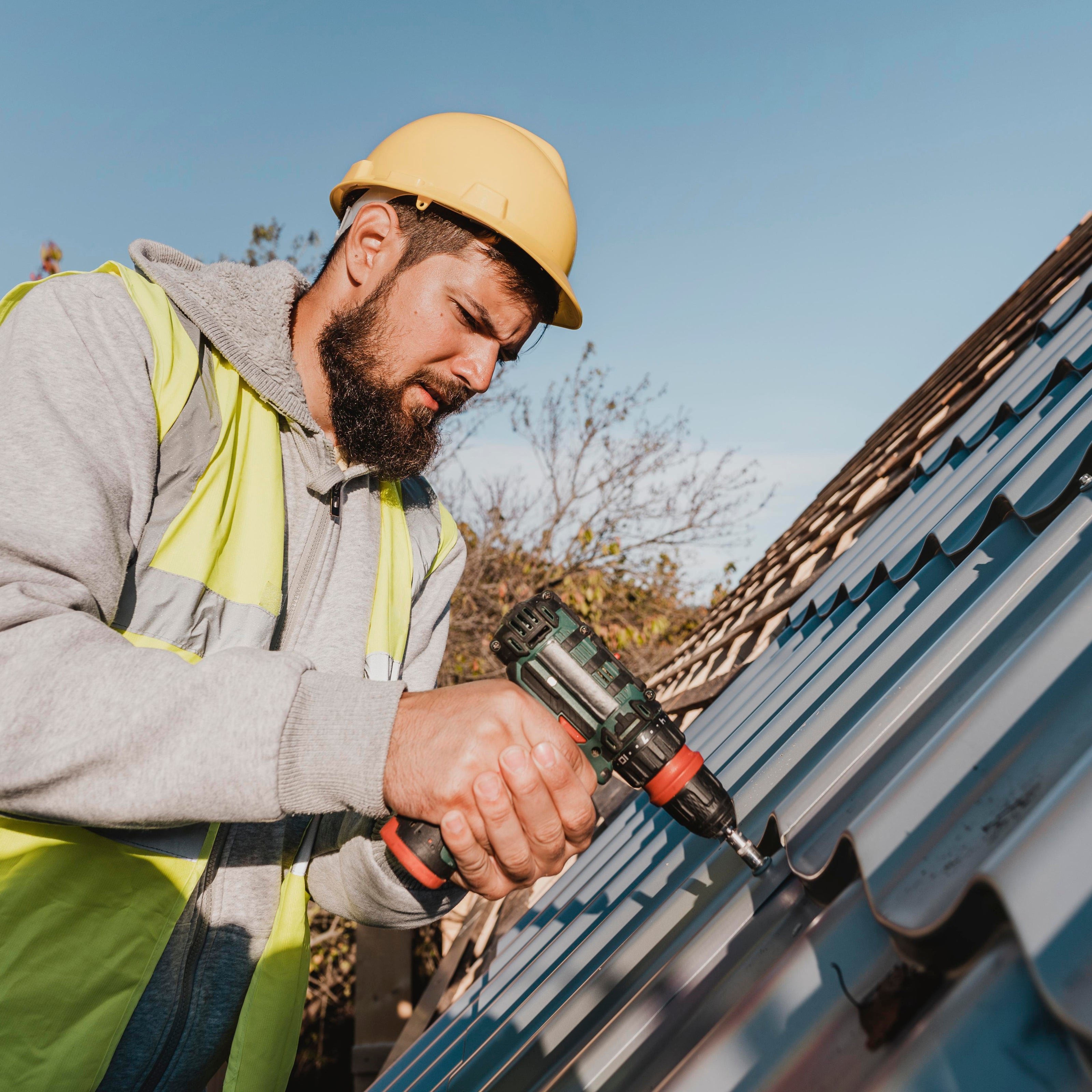 Person working on a roof with a drill, wearing safety gear including a hard hat and high-visibility jacket.